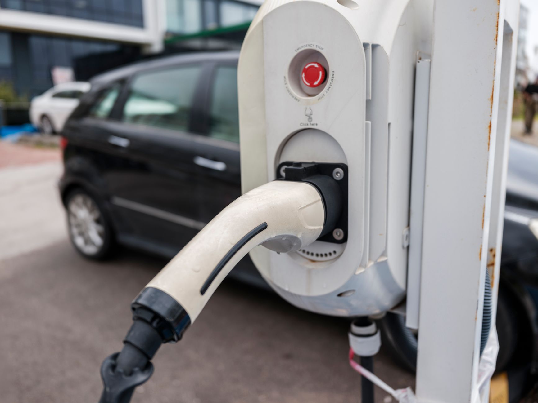 Borne de recharge blanche avec connecteur au premier plan devant une voiture noire garée en extérieur.
