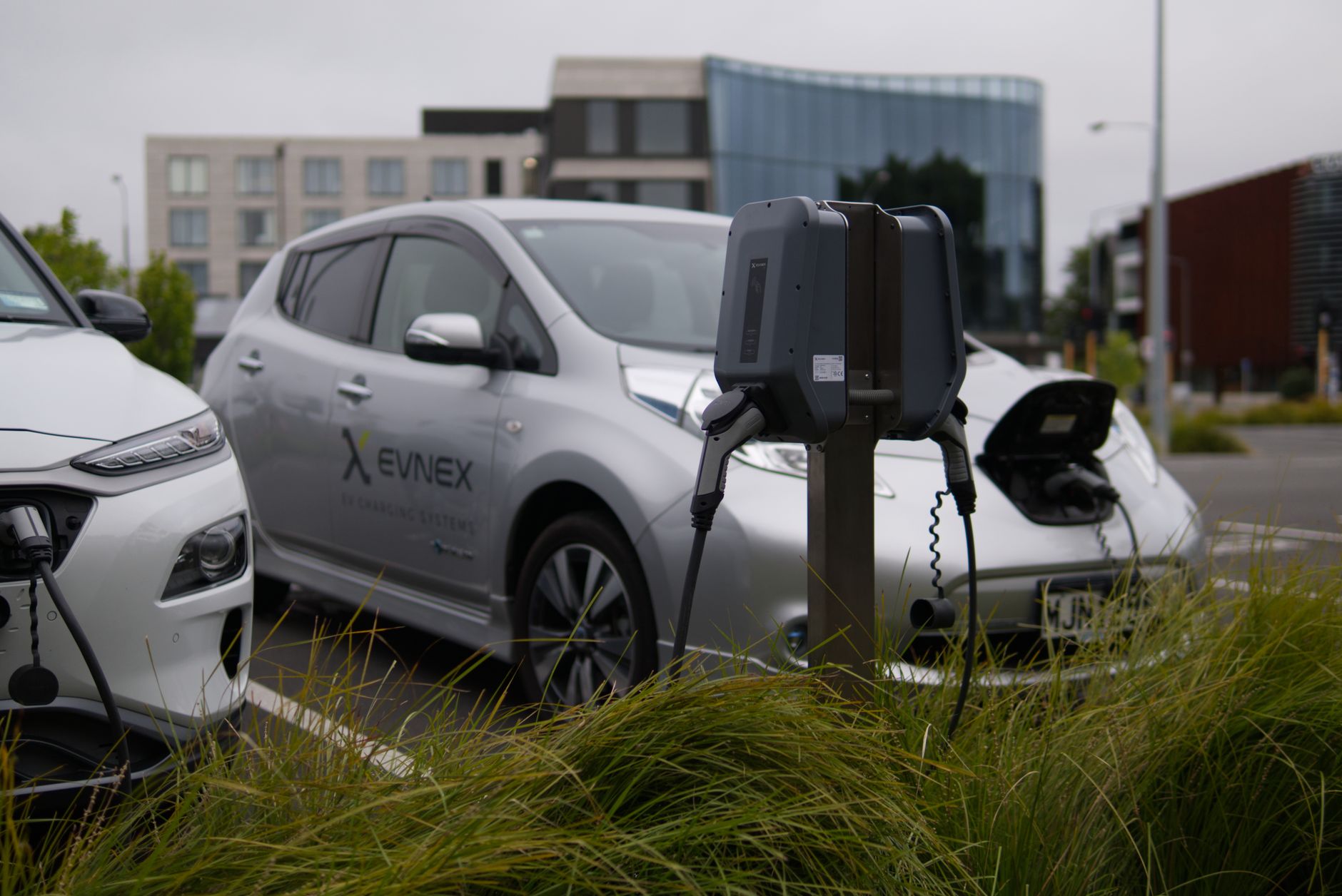 Borne de recharge avec câbles branchés sur deux voitures électriques garées sur un parking, devant des immeubles modernes.
