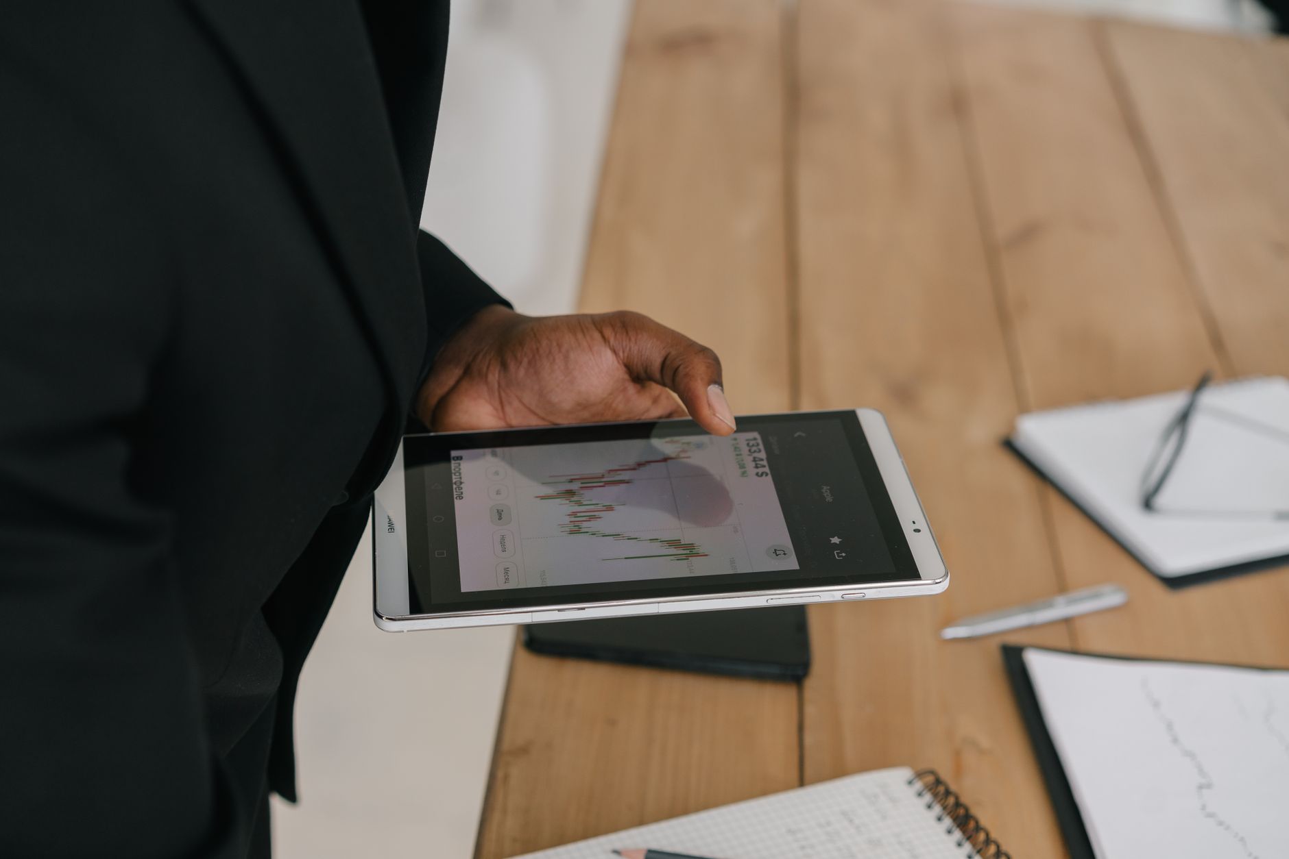 Une personne en veste noire consulte un graphique de données sur une tablette, près d’une table en bois avec carnets, stylo et lunettes.